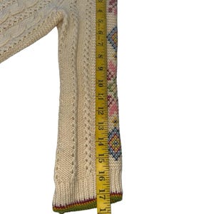 Op de afbeelding: Cr&egrave;mekleurige trui met ingewikkeld kabelbreiwerk en geborduurde bloemen- en geometrische patronen op de mouw. De mouw is ongeveer 48 cm lang. De trui heeft een groene, rode en gele bies.