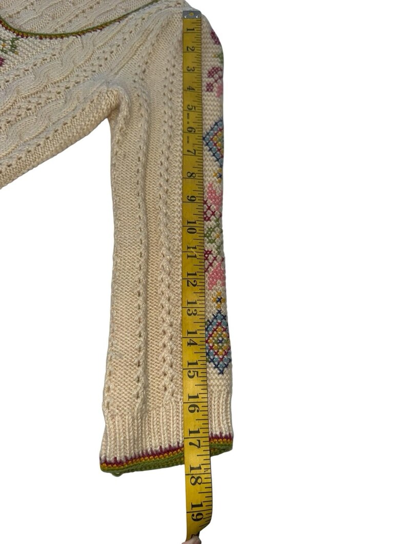 Op de afbeelding: Cr&egrave;mekleurige trui met ingewikkeld kabelbreiwerk en geborduurde bloemen- en geometrische patronen op de mouw. De mouw is ongeveer 48 cm lang. De trui heeft een groene, rode en gele bies.