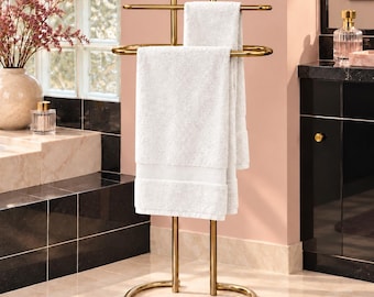 Può includere: Un portasciugamani da terra in metallo color oro con base circolare e due barre orizzontali. Un asciugamano rettangolare bianco è drappeggiato sulla barra superiore e ricade su quella inferiore, mostrando un design pulito e funzionale.