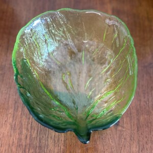 Op de afbeelding: Een decoratieve glazen kom in de vorm van een blad. De kom is doorschijnend groen met donkergroene accenten langs de randen en nerven. De kom staat op een houten oppervlak.