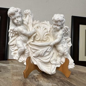 Könnte beinhalten: Eine weiße Cherubinskulptur mit zwei geflügelten Figuren, die einen drapierten Stoff um ein dekoratives Becken halten. Die Cherubim haben lockiges Haar und stehen auf einem Holzständer. Die Skulptur ist ein dekoratives Stück.