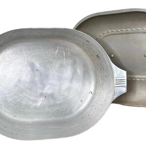 May include: An oval aluminium casserole dish with a matching lid. The dish has two handles with a ribbed design. The lid is slightly smaller and has a raised oval shape in the centre. The dish and lid are both silver in colour.