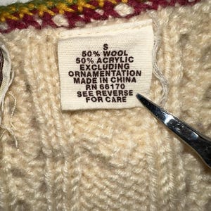 Op de afbeelding: Close-up van een cr&egrave;mekleurige gebreide trui met een label. Het label luidt "50% Wol, 50% Acryl, Exclusief Ornamentatie, Made in China, RN 66170, Zie achterkant voor verzorging." De trui heeft een decoratieve afwerking.