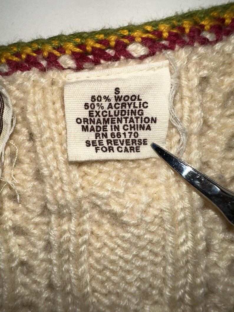 Op de afbeelding: Close-up van een cr&egrave;mekleurige gebreide trui met een label. Het label luidt "50% Wol, 50% Acryl, Exclusief Ornamentatie, Made in China, RN 66170, Zie achterkant voor verzorging." De trui heeft een decoratieve afwerking.