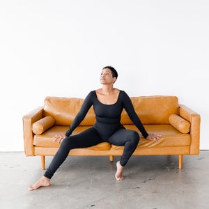 May include: A person wearing a black long-sleeved jumpsuit sits on a brown leather couch with their legs spread out. The person is looking up and has short black hair.