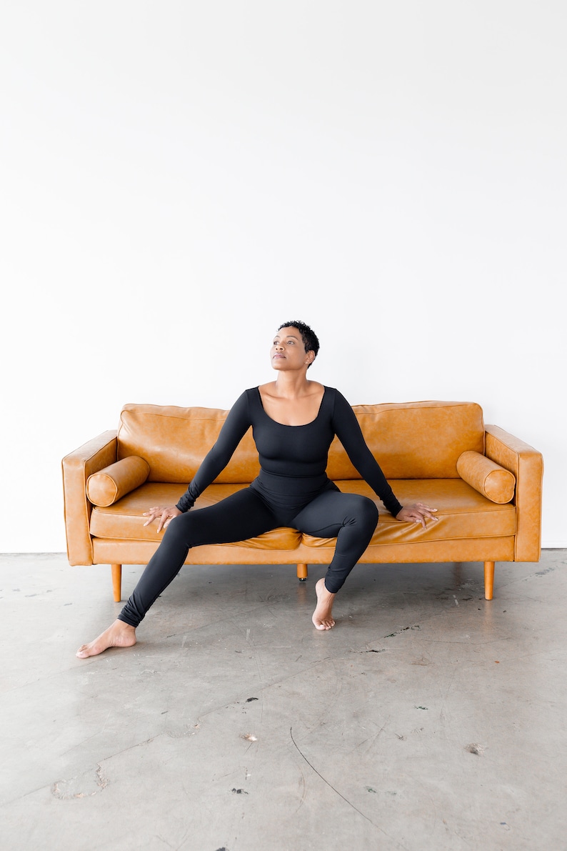 May include: A person wearing a black long-sleeved jumpsuit sits on a brown leather couch with their legs spread out. The person is looking up and has short black hair.