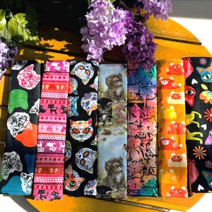 Könnte beinhalten: Acht bunte Stoffstreifen mit verschiedenen Katzendesigns. Die Streifen sind horizontal angeordnet und zeigen verschiedene Muster, darunter Schädel, Blumen und geometrische Formen.
