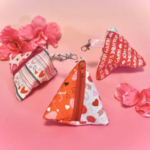 May include: Three small triangular zippered pouches with Valentine's Day themed fabric. The pouches are red, white, and pink with hearts and the text "Happy Valentine's Day".