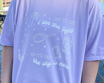 Puede incluir: Una camiseta de color lavanda con un diseño gráfico blanco que incluye una sartén, una flor y el texto "AND AT LAST i see the light AND IT'S LIKE the sky is new".