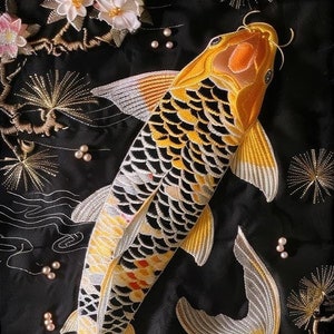 May include: Two embroidered koi fish swimming in a black fabric with gold and white floral designs. The fish are yellow, orange, and black with white accents.