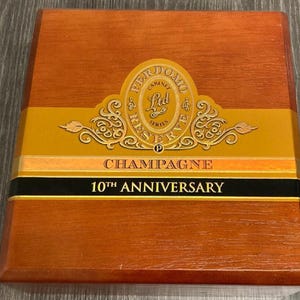 May include: A wooden box with a light brown finish. The box features a gold and black label with the words "PERDOMO CABINET RESERVE" and "CHAMPAGNE 10TH ANNIVERSARY" in gold lettering.