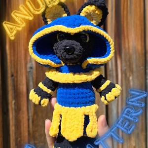 May include: A blue, yellow, and black crochet Anubis doll. The doll features a hood, ears, and a skirt-like design. The image includes the text "ANUBIS" and "PDF PATTERN".