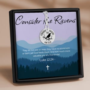 Puede incluir: Collar con colgante plateado con un dije redondo que presenta una ilustración de un cuervo y el texto "Consider the Ravens". El collar se exhibe en una caja negra con un fondo que incluye texto de Lucas 12:24 y una cruz.