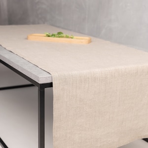 Könnte beinhalten: Ein beiger Tischläufer aus Leinen auf einem schwarzen Metalltisch mit einem Holztablett darauf. Der Tischläufer ist aus natürlichem Leinen gefertigt und hat eine dezente Textur.