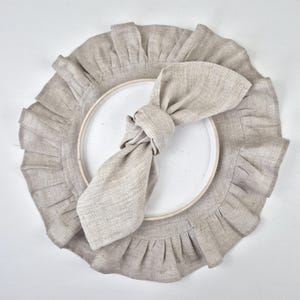 Peut inclure: Un set de table en lin beige avec un bord à volants, une assiette blanche et une serviette nouée assortie. Le set de table et la serviette sont en tissu texturé. Les articles sont disposés sur une surface blanche, créant une table simple et élégante.