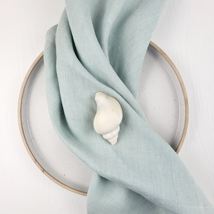 Pode incluir: Um guardanapo de linho azul claro está dobrado em um prato de cerâmica branco com uma concha branca no centro.