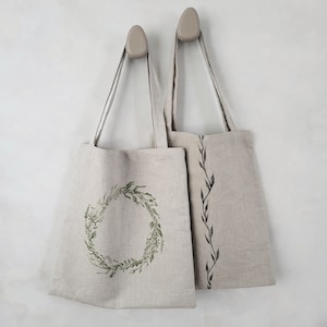 Puede incluir: Dos bolsas de mano beige colgadas en una pared. Una bolsa presenta un diseño de corona de hojas verdes, mientras que la otra tiene un patrón de hojas vertical. Ambas bolsas tienen correas largas y delgadas y están hechas de una tela de aspecto natural.