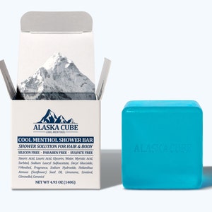 May include: A blue shower bar with the text "ALASKA CUBE" in white. The bar is in a white box with the text "COOL MENTHOL SHOWER BAR" and "SHOWER SOLUTION FOR HAIR & BODY" in black. The box also lists the ingredients and net weight of the bar.