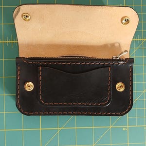 Könnte beinhalten: Eine schwarze Ledergeldbörse mit Reißverschluss und Klappe. Die Geldbörse hat ein Fach innen und zwei goldene Druckknöpfe außen.