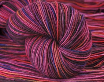 May include: A skein of yarn in a multi-colored pattern of deep red, pink, purple, and black. The yarn is wound into a ball shape.