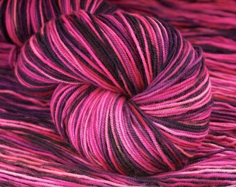May include: A skein of yarn with pink, purple, and black stripes. The yarn is wound into a ball shape and is lying on a surface of the same yarn.
