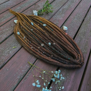 Può includere: Dreadlock sintetiche marroni e bionde con perline e conchiglie, su una superficie di legno con fiori bianchi e foglie verdi.