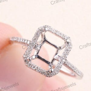 Könnte beinhalten: Ein silberner Ring mit quadratischem Halo-Design. Der Ring hat eine zentrale quadratische Fassung, die von kleinen, funkelnden Steinen umgeben ist. Das Band ist ebenfalls mit kleineren Steinen verziert. Der Text "Craftsjewels" ist sichtbar.