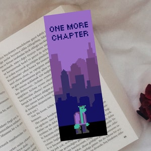 Peut inclure: Un marque-page pixelisé violet et bleu avec le texte "ONE MORE CHAPTER" et une ville en arrière-plan. Un personnage de monstre vert se tient devant la ville.