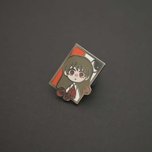 May include: A silver enamel pin featuring a cartoon character with brown hair, red eyes, and a red bow tie. The character is wearing a white shirt and is surrounded by red roses. The pin is set against a red and white background.