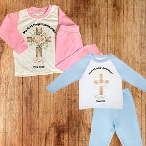 May include: Two sets of children's pajamas. One set is pink and white with a cross design and the text "My first Holy Communion" and "Lilly May 2026". The other set is blue and white with the text "My first Holy Communion" and "Cathal May 2026".