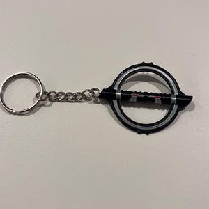 May include: A black and silver keychain with a circular design and a small lightsaber-shaped charm. The keychain has a silver chain and a silver ring.