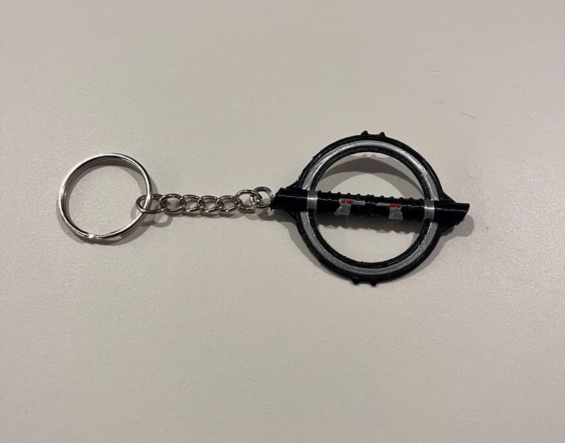 May include: A black and silver keychain with a circular design and a small lightsaber-shaped charm. The keychain has a silver chain and a silver ring.
