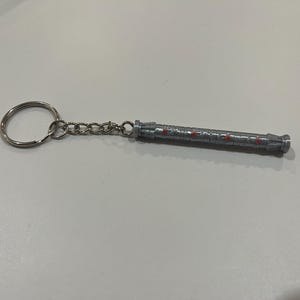 May include: A silver keychain with a detailed, cylindrical charm. The charm has red accents and a ring at the end.