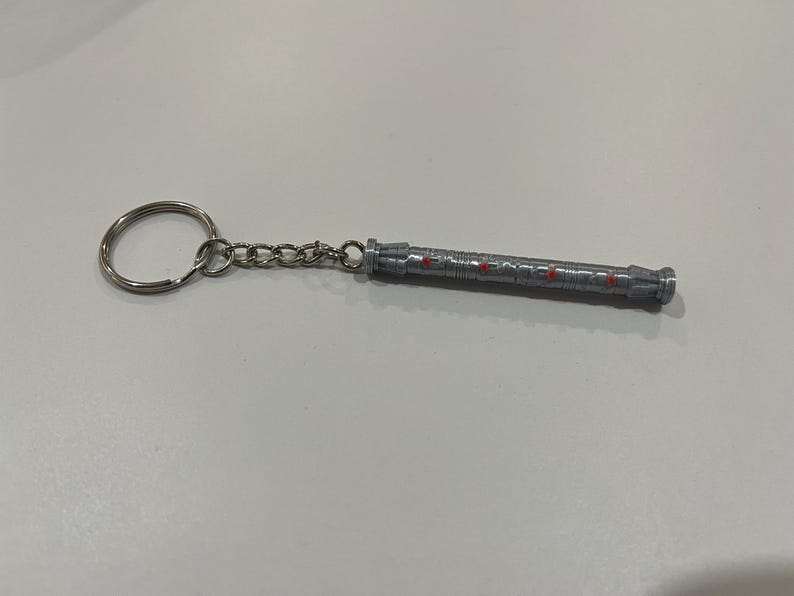 May include: A silver keychain with a detailed, cylindrical charm. The charm has red accents and a ring at the end.