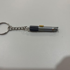 May include: A silver lightsaber keychain with a black handle and a yellow button. The keychain is attached to a silver key ring.