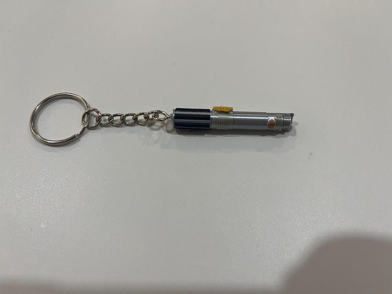 May include: A silver lightsaber keychain with a black handle and a yellow button. The keychain is attached to a silver key ring.