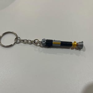 May include: A silver keychain with a black and silver lightsaber charm. The lightsaber has a gold accent.