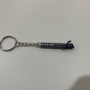 May include: A silver keychain with a black lightsaber charm. The lightsaber is detailed with silver stripes and a black handle.