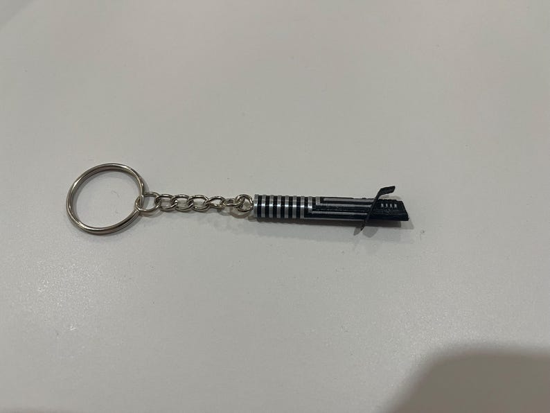 May include: A silver keychain with a black lightsaber charm. The lightsaber is detailed with silver stripes and a black handle.