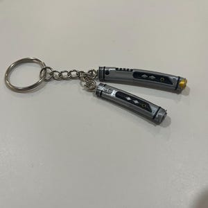 May include: A silver keychain with two small, silver lightsaber-shaped charms. The charms have black details and a gold accent.