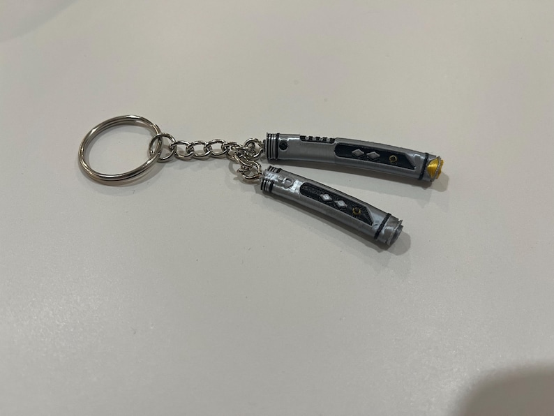 May include: A silver keychain with two small, silver lightsaber-shaped charms. The charms have black details and a gold accent.