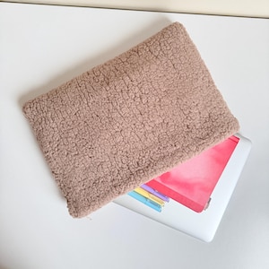 May include: A tan, fuzzy laptop sleeve rests on top of a white laptop. The sleeve has a rectangular shape and a soft, textured appearance. The laptop screen is partially visible, displaying a pink and colorful design.