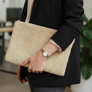 May include: A beige, rectangular, fluffy laptop sleeve is held against a black blazer. The sleeve has a textured, plush appearance. A silver watch is worn on the wrist. The background is blurred, suggesting an office or indoor setting.
