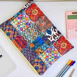 Puede incluir: Una funda para portátil de patchwork colorida con cremallera amarilla. La funda presenta una variedad de cuadrados de tela estampada en rojo, azul, naranja y verde. Se ve una calculadora rosa y bolígrafos.