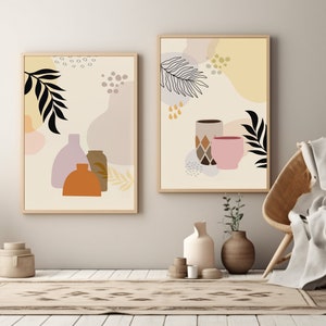 May include: Two framed abstract art prints with a beige background. The print on the left features a black leaf, a pink and orange abstract shape, and three vases in shades of purple, brown, and orange. The print on the right features a black leaf, a pink and orange abstract shape, and two cups in shades of brown and pink.