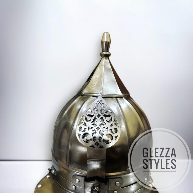May include: A silver-coloured, medieval-style helmet with a pointed top and decorative filigree. The helmet has a rounded dome shape and is made of metal. The text "GLEZZA STYLES" is visible in a white circle.