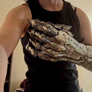 Op de afbeelding: Een metalen handschoen met gearticuleerde vingerplaten met puntige uiteinden en ingewikkelde gegraveerde patronen. Het pantserstuk bedekt de hand en pols, opgebouwd uit gesegmenteerde zilverkleurige metalen platen vastgezet met klinknagels.
