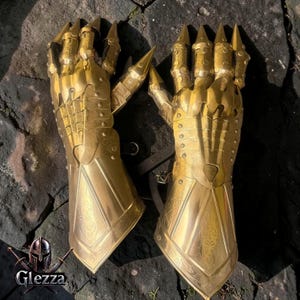 Op de afbeelding: Een paar goudkleurige metalen handschoenen met gearticuleerde vingerplaten met puntige uiteinden, overlappende polssecties en ingewikkelde gegraveerde patronen op de onderarmmanchetten, vergezeld van een klein logo met de tekst Glezza.