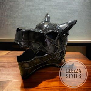 May include: A black, stylized helmet shaped like a wolf's head, with pointed ears and a detailed, geometric design. The helmet is made of a shiny material and sits on a wooden surface. The text "GLEZZA STYLES" is in a circle on the bottom right.