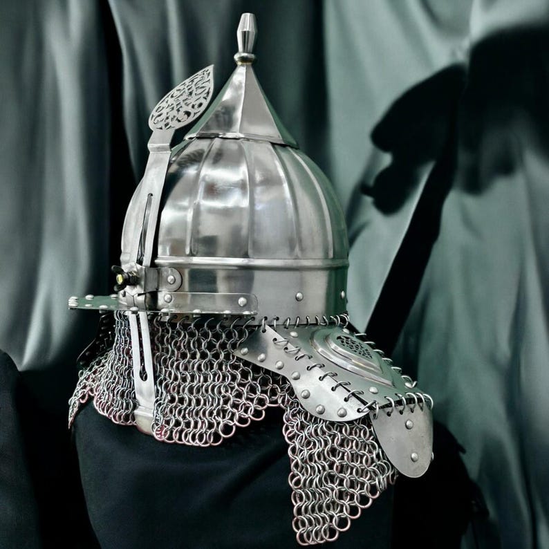 May include: A silver medieval helmet with a rounded crown and a pointed top. It has a face guard, decorative details, and a chainmail neck protector. The background is a muted grey.